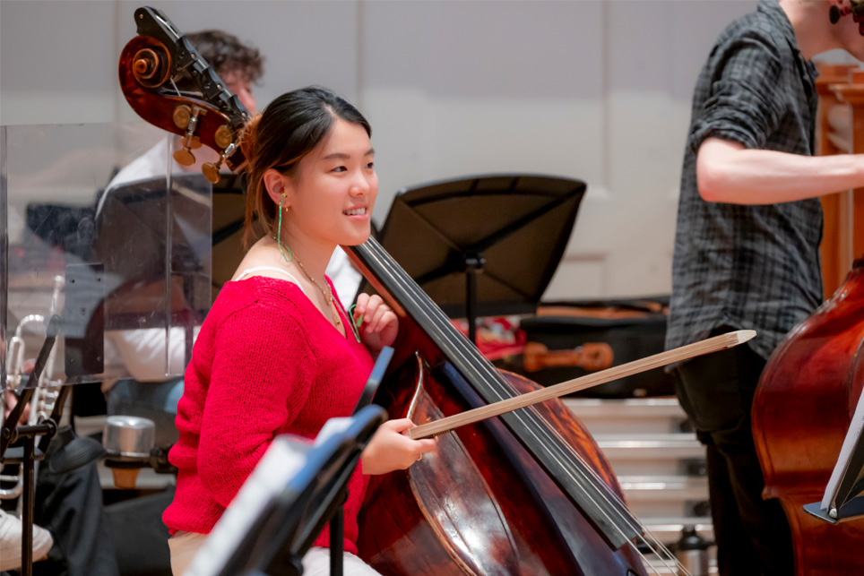 A female double bass player, holding her bow up and her double bass leaning against her, chatting to other students, sitting down in an orchestra rehearsal.